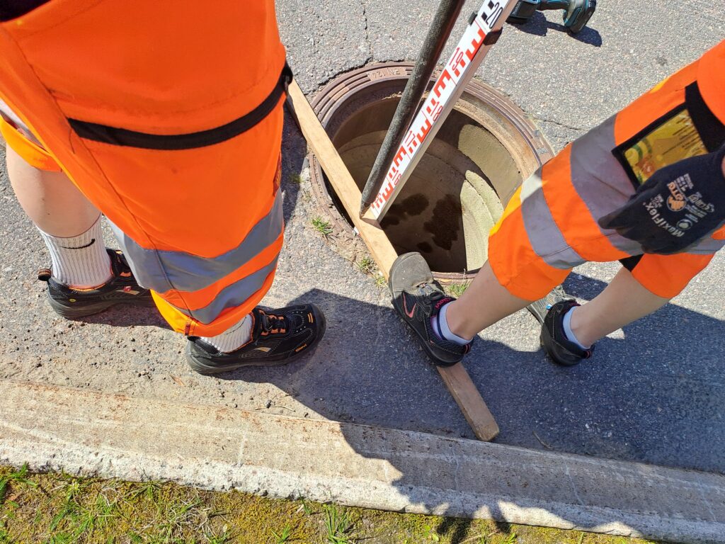 Kaksi henkilöä oransseissa työvaatteissa, vain jalat näkyvät. Kaivon kansi poissa ja mittaustyö käynnissä.