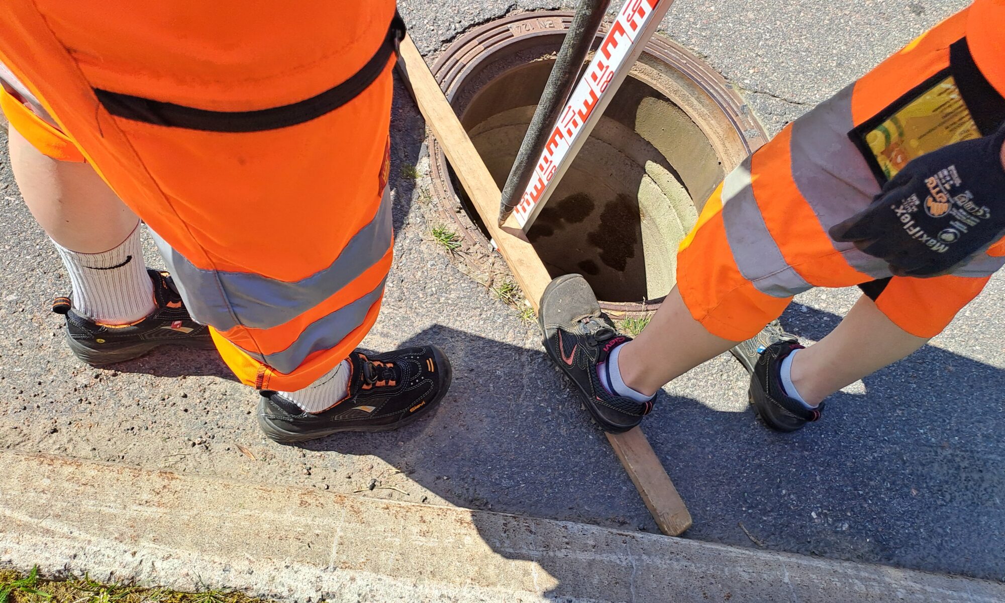 Kaksi henkilöä oransseissa työvaatteissa, vain jalat näkyvät. Kaivon kansi poissa ja mittaustyö käynnissä.