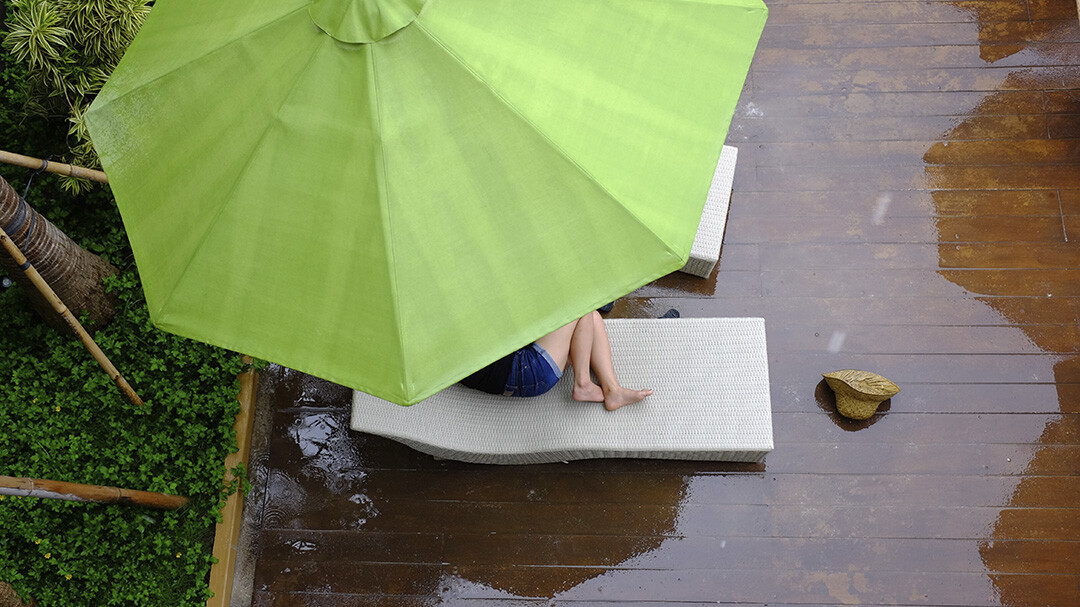 Nainen lepäilee terassilla ison aurinkovarjon alla suojassa vesisateelta.