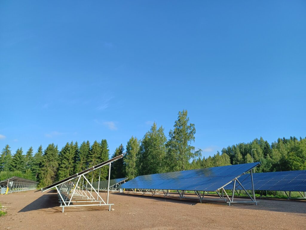 Maa-asenteisia aurinkopaneeleita hiekkakentällä aurinkoisena päivänä, takana puita ja sininen taivas.