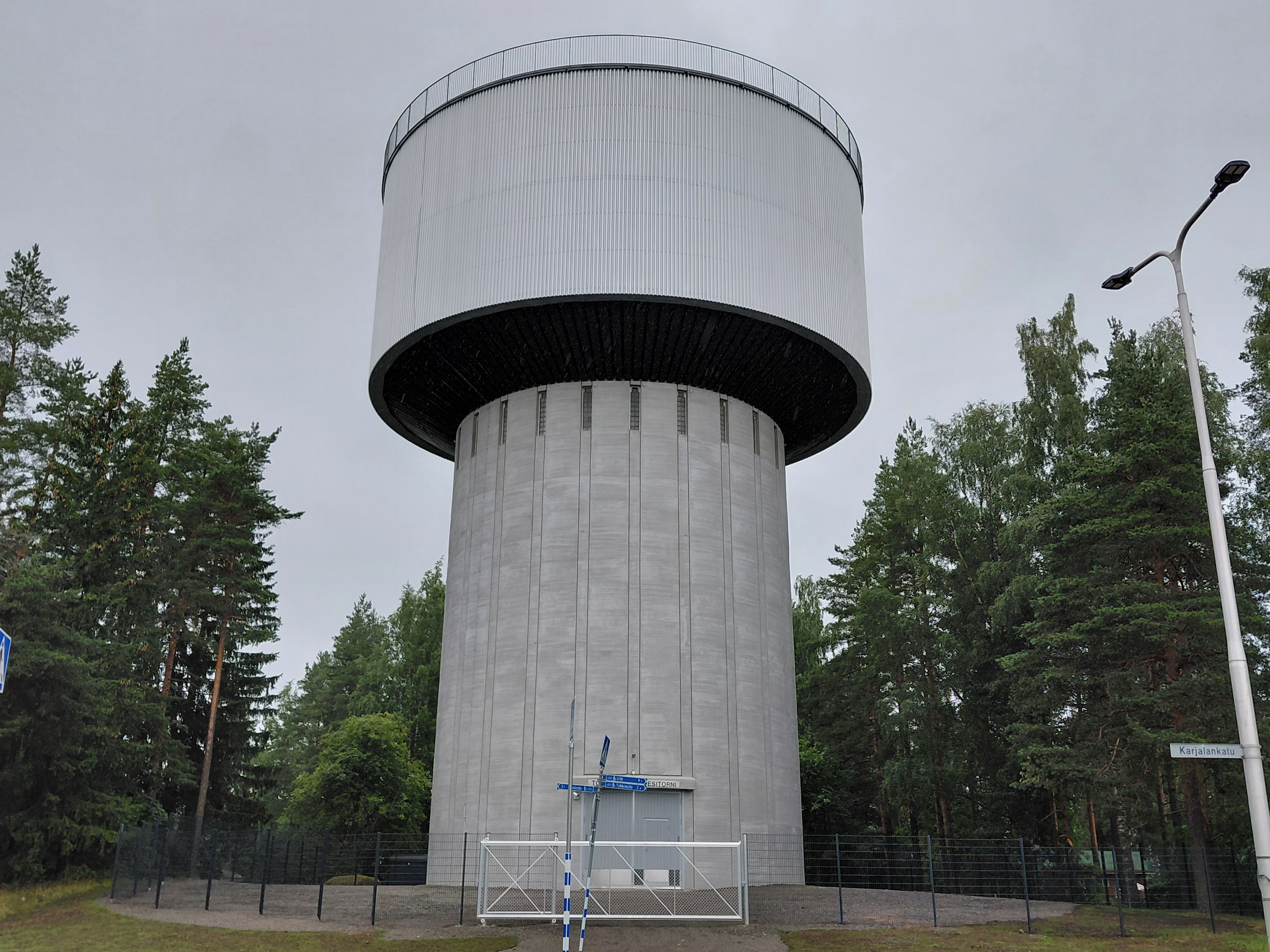 Harmaa vesitorni puuston keskellä. Tornin piha aidattu.