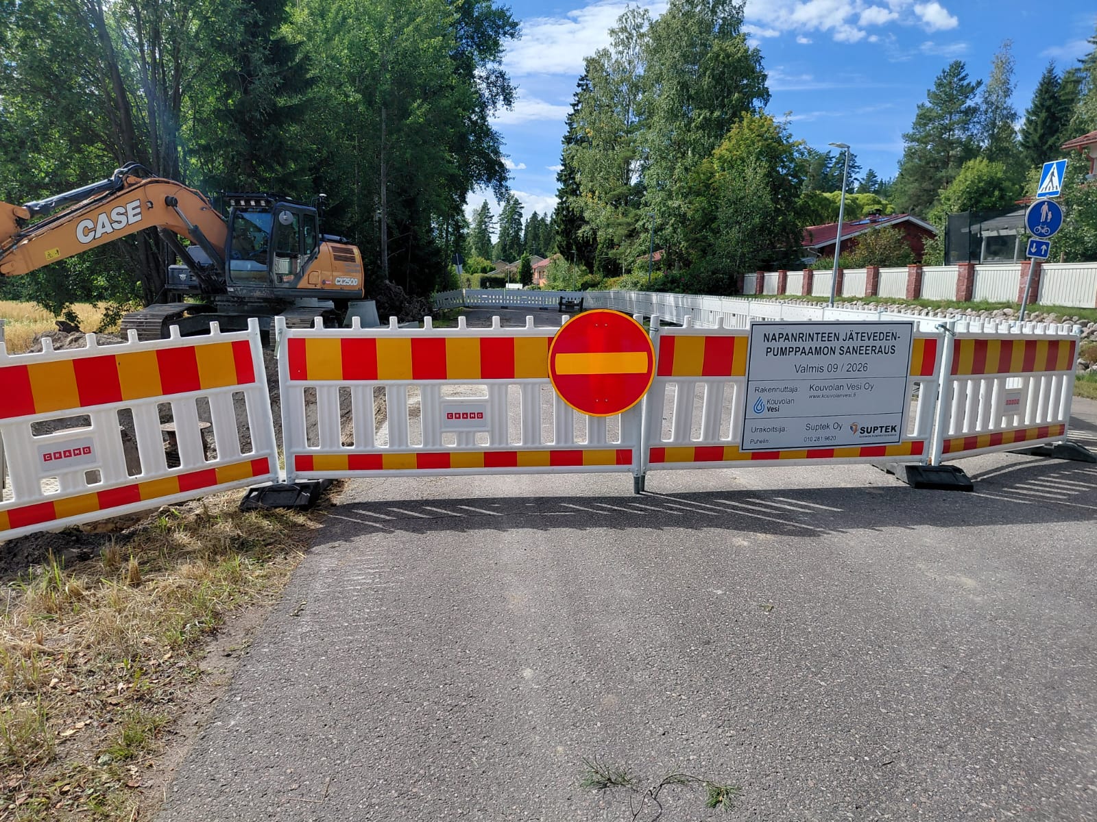 Ajotiellä rajattu työmaa-alue valkoisilla työmaa-aidoilla kesällä. Kielletty ajosuunta.merkki aidassa. Aidan sisäpuolella kaivinkone.
